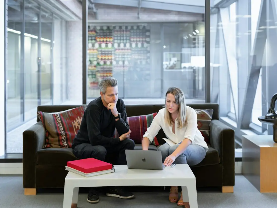 A man and a woman working on a laptop on a sofa in a bright office